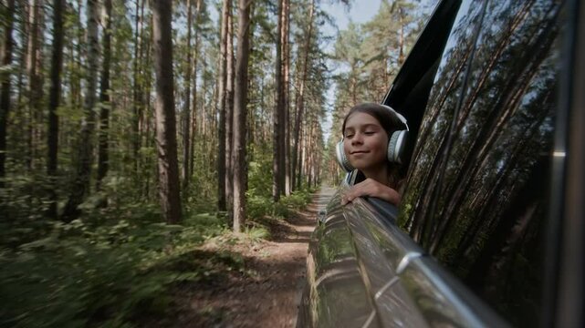 Young carefree boy with in headphones leaning out of car window, looking at scenic forest road with tall trees and lush greenery around, enjoying summer outing