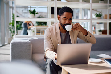 Black corporate manager drinking coffee while reading email on laptop.