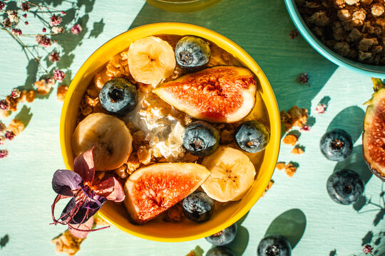 Healthy breakfast, figs, bananas and blueberries in yellow and blue bowls with yogurt and granola on a light table. View from above.