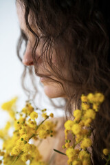 Naklejka premium A delicate portrait of a woman with wavy hair, partially hidden by yellow mimosa flowers. Soft lighting and a dreamy atmosphere evoke feelings of spring, femininity, and serenity.