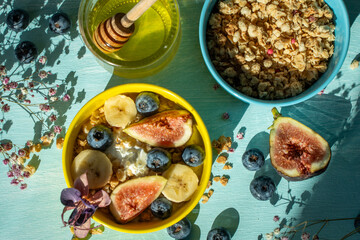 Healthy breakfast, figs, bananas and blueberries in yellow and blue bowls with yogurt and granola on a light table. View from above.