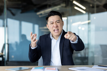 Portrait of angry young Asian man in business suit sitting at desk in office, talking and pointing aggressively at camera