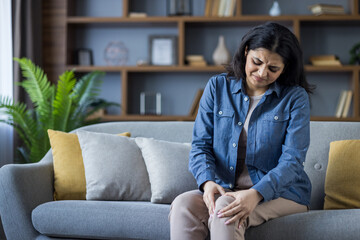A young Muslim woman sits on the couch at home and holds her leg with her hands, feeling severe pain in her knee and bones