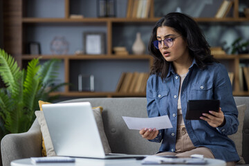 Worried young Muslim woman sitting at home on the couch behind a laptop, looking at the screen in...
