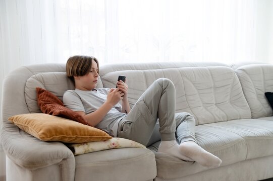 Teenager works on homework while relaxing on a couch in a cozy living room during the afternoon. Full length portrait of young teenage boy in hand smartphone while sitting on sofa
