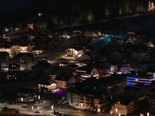 Obraz premium A nighttime scene of Ischgl ski resort in Austria, featuring glowing alpine style buildings, illuminated slopes, and a snow covered hillside with a forested backdrop.