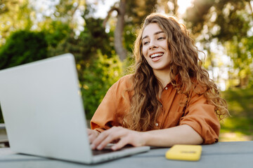 Happy woman sits in park with laptop. Portrait of a young freelance woman working outdoors. Online, technology concept