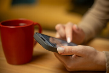 A person is engaged in using a smartphone while seated at a wooden table, with a steaming red mug nearby. The indoor atmosphere is warm and inviting.