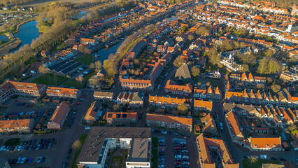 An aerial view of a European town with red-roofed houses, narrow streets, and a canal winding through the landscape. The warm sunlight highlights the architecture and surrounding greenery.