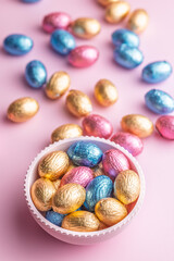 Chocolate easter eggs wrapped in multi colored foil in bowl on pink background.