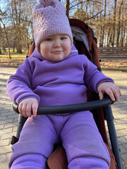 Child in Purple Winter Outfit Enjoys Sunny Day in Park While Sitting in Stroller
