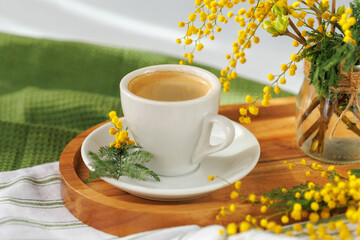 A cup of coffee placed on a wooden tray, with soft sunlight highlighting the vibrant mimosa flowers, perfect for a relaxing morning.
