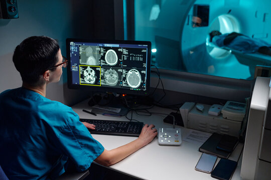Medical professional reviewing patient's MRI images on computer monitor with patient in MRI machine in background. Hospital equipment and technology setup visible