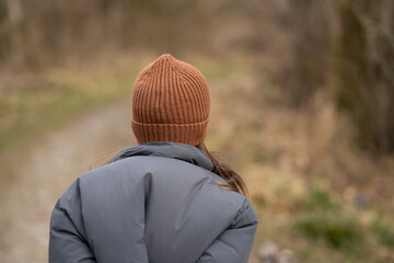 A girl with her back to the camera wearing a brown hat and jacket outside on a path in the woods....