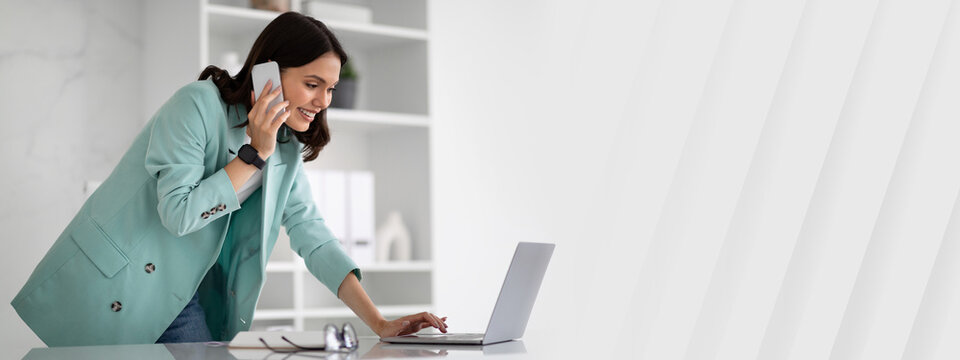 Busy happy young european woman in suit typing on laptop, calling by phone in modern office interior. Businesswoman, multitasking and business, work, lifestyle, client communication