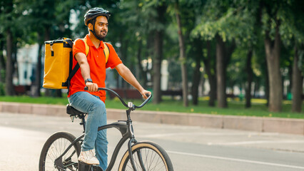 Obraz premium Modern work and delivery on eco transport. Young friendly indian man in uniform, protective gear with backpack riding on bicycle on sidewalk down the street, side view, panorama, empty space, outdoor