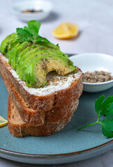 Avocado toast with cream cheese on rustic sourdough bread, sprinkled with black pepper and fresh parsley. Served on a ceramic plate with lemon wedges and spices. Close up.
