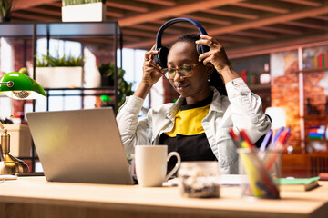 Freelancer in home office putting headphones on, using laptop device to work on tasks. African american remote employee preparing to listen music while using notebook to do job online
