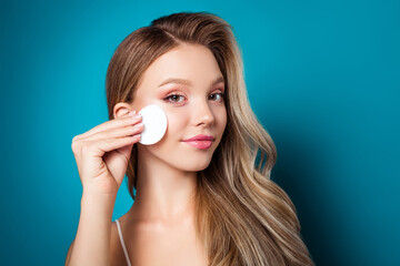 Young blonde woman practicing skincare routine using cotton pad against blue background