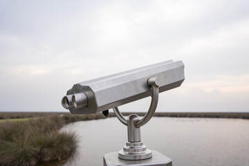Metallic binocular overlooking marsh with water and path under overcast sky. Closeup of outdoor coin operated silver binocular viewer for tourists. Distant landscape. Telescope, travel, nature concept