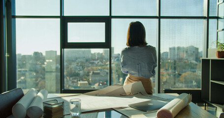 An architect standing by a window overlooking a cityscape, reflecting on project plans with...