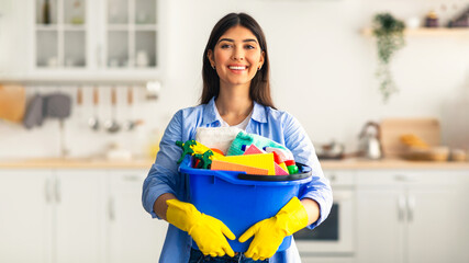 A young woman in her twenties stands in a bright kitchen, holding a blue bucket filled with various...