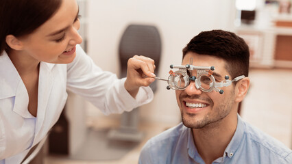 A smiling man is undergoing an eye examination in a well-lit clinic. An optometrist adjusts his...