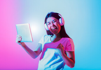 A young Asian woman listens to music through headphones while holding a tablet. She is visibly...