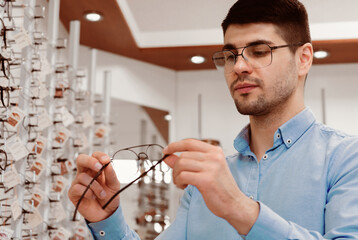 A young man is carefully looking at a pair of eyeglasses inside an optical store, considering...