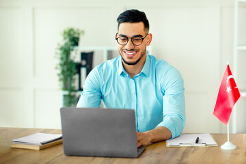 Fototapeta premium A young Arab entrepreneur is focused on his laptop while sitting at a desk in a contemporary office. His expression shows determination as he enhances his professional skills.