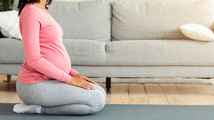 Fototapeta premium Side view of pregnant black woman sitting on yoga mat, getting ready before morning exercising, home interior, free space, cropped. Unrecognizable african expecting woman meditating alone at home