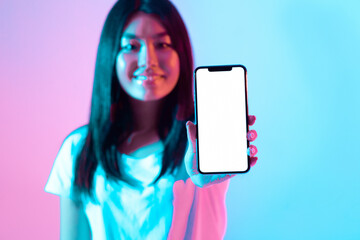 A young woman holds up a smartphone in front of her with a blank screen. The vibrant blue and pink...