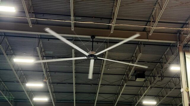 A large, industrial ceiling fan with five white blades spins in a high-ceilinged space, featuring exposed metal beams and rectangular light panels