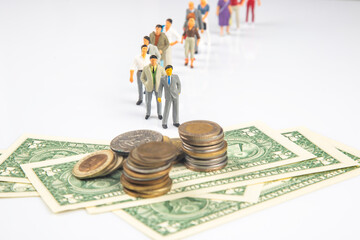 Miniature people forming a queue against a blurred background, paired with piles of coins and...