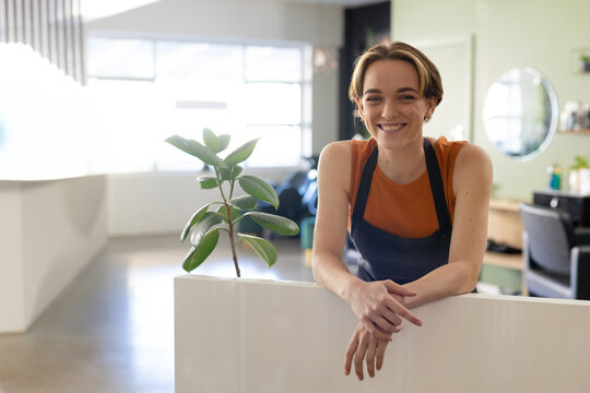 Smiling barber leaning on counter in modern salon, welcoming customers warmly, copy space