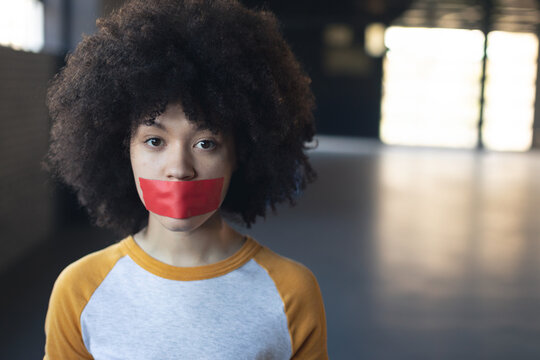 Young person with red tape on mouth standing in spacious warehouse, looking serious, copy space