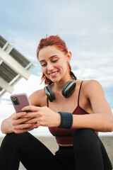 Verttical. Happy young woman enjoying smartphone while sitting outdoors with headphones, engaged in...