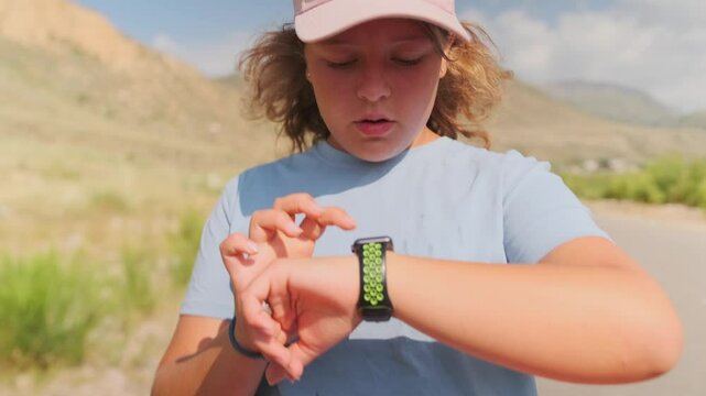 After a long run through greenery and mountains, a full figured teenage girl reaches the finish line. Checking the timer on her sports watch, she wipes sweat from her brow, resting after her efforts.