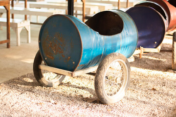 A whimsical toy car constructed from repurposed metal barrels and wheels, standing on a gravel surface under bright sunlight. Evokes a sense of childhood creativity, DIY projects, and rustic charm.