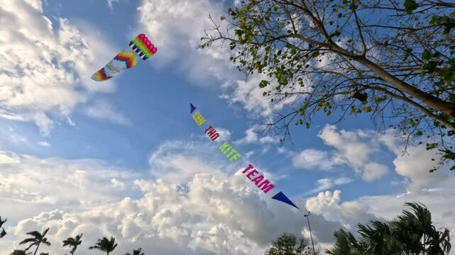 A colorful kite with a banner soars high in the sky against blue clouds and a tree with green leaves.
