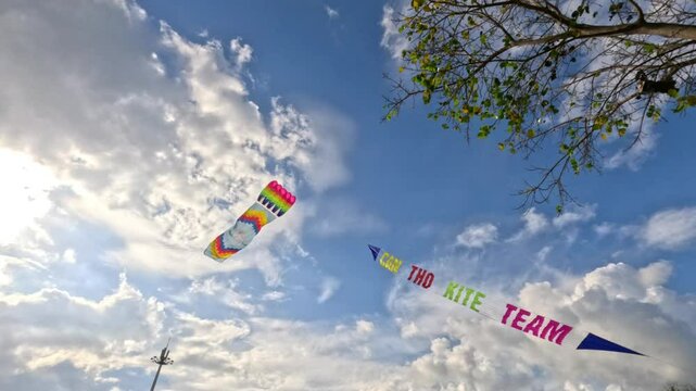 A colorful kite with a banner soars high in the sky against blue clouds and a tree with green leaves.