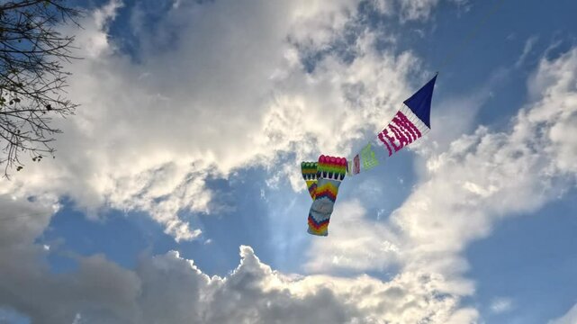 Colorful kites with a banner soars high in the sky against blue clouds and a tree with green leaves.