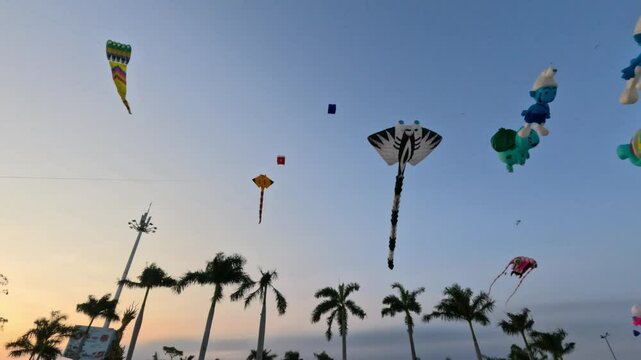 A variety of colorful kites, shaped like animals and cartoon characters, soar high in the sky against the backdrop of palm trees and a sunset lit horizon in Can Tho, Vietnam - Feb 25, 2025.