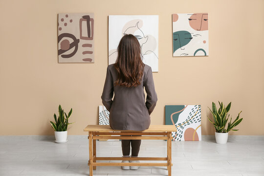 Young woman sitting on bench near paintings on beige wall in art gallery, back view
