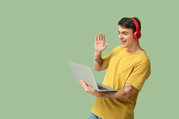 Handsome young man with headphones and laptop waving hand on green background