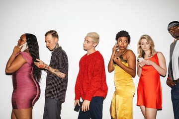 A diverse group of people standing in line, using phones. Includes men and women of various ethnicities, each engaged with their mobile devices. Party people waiting in line at club.