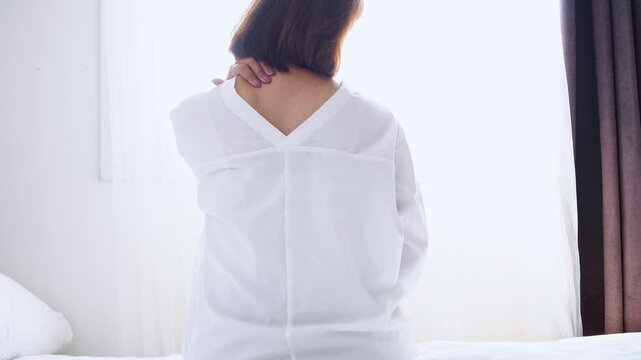 Asian woman sitting in bed feeling neck and shoulder, upper back muscle pain after wake up due to sleep wrong position 