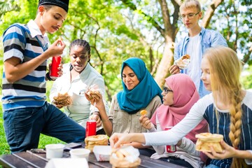 Group of diverse young people enjoying a picnic in a park, sharing food and drinks, sitting on grass, smiling and interacting in a relaxed outdoor setting. Diverse students eating in a park.