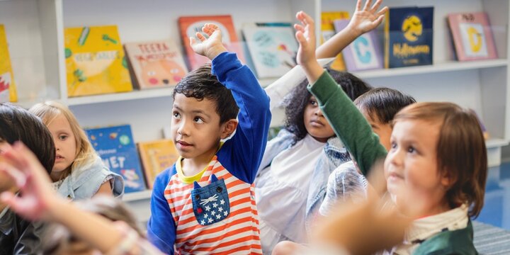 Children in a classroom raising hands, eager to participate. Diverse group of kids, engaged in learning. Classroom setting with books in the background. Little diverse kids raising hands in classroom.