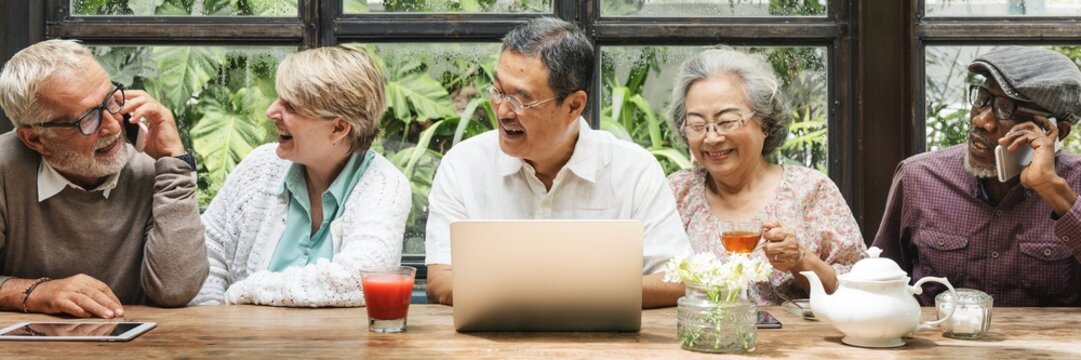 Diverse group of seniors enjoying conversation. Elderly friends, men and women, chatting and laughing. Seniors bonding over drinks and technology. Senior people using digital devices and social media. - Powered by Adobe